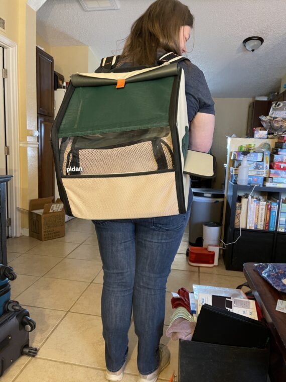 A woman, facing away from the camera, is wearing a large backpack-cat carrier combo. There is a small orange-and-white cat inside the carrier, though he is hard to see.
